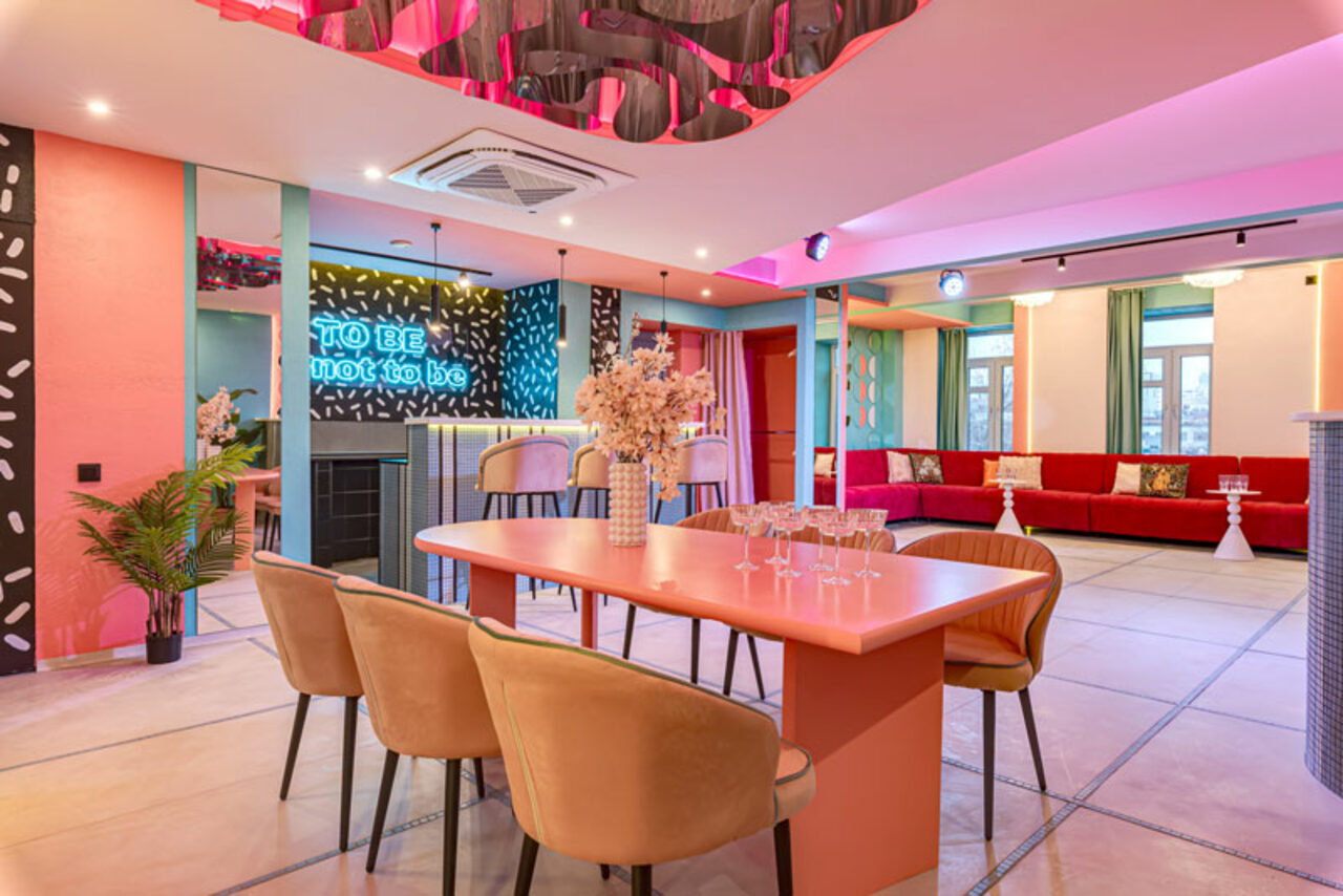 Арендовать Лофт Шекспир. Стильный дизайнерский зал. Просторное помещение с ярким интерьером: длинный стол с несколькими стульями, диван вдоль стены, надпись на стене "TO BE not to be". Разноцветные стены и потолок, зонированное освещение, декоративные элементы, кондиционер на потолке.