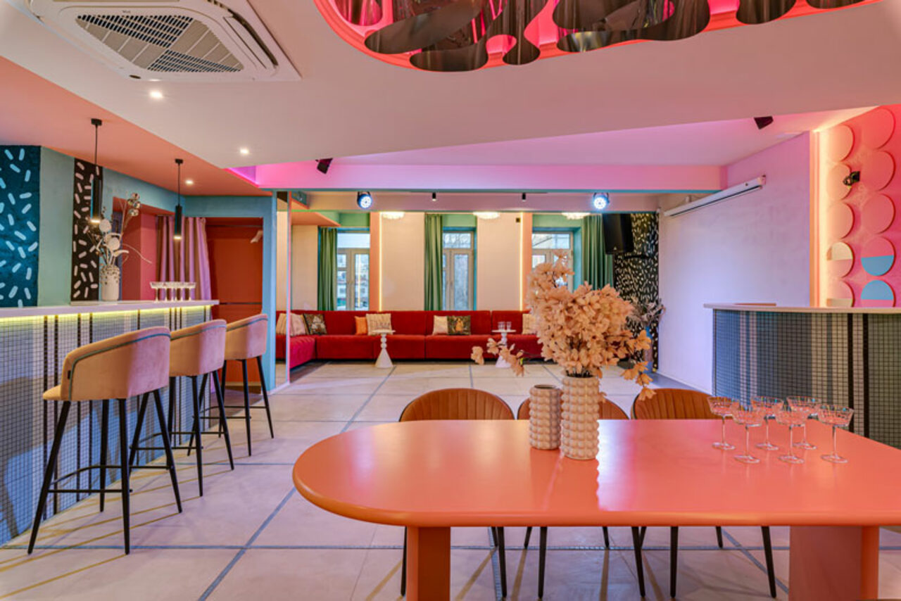 Арендовать Лофт Шекспир. Стильный дизайнерский зал. Просторное помещение с барной стойкой и высокими стульями, обеденным столом и стульями, декорированное цветными подсветками. Цветовая гамма включает розовый, голубой и бежевый. Стены украшены узорами. На столе бокалы и ваза с цветами.