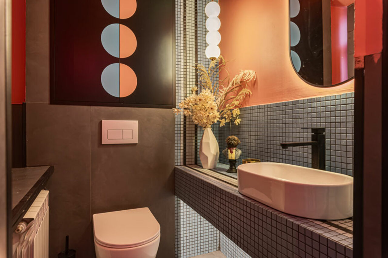 Арендовать Лофт Шекспир. Стильный дизайнерский зал. Современная ванная комната. Плитка: мелкая мозаика серая. Умывальник белый, на черной тумбе. Зеркало овальное с подсветкой. Унитаз белый, встроенный. Декор: ваза с сухоцветами. На стене художественный постер с геометрическими фигурами.