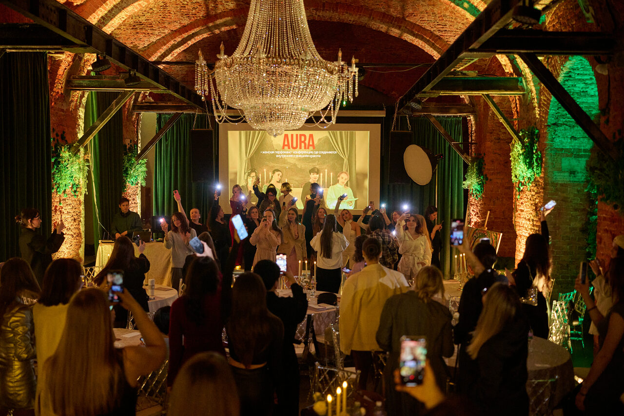 Арендовать Arsenal Loft(Арсенал Лофт). Арочный зал с кирпичными стенами, большим кристальным люстрой и деревянными балками. Пространство заполнено людьми, на сцене группа. На заднем плане светящийся экран с надписью "AURA". Столы с белыми скатертями и прозрачными стульями.