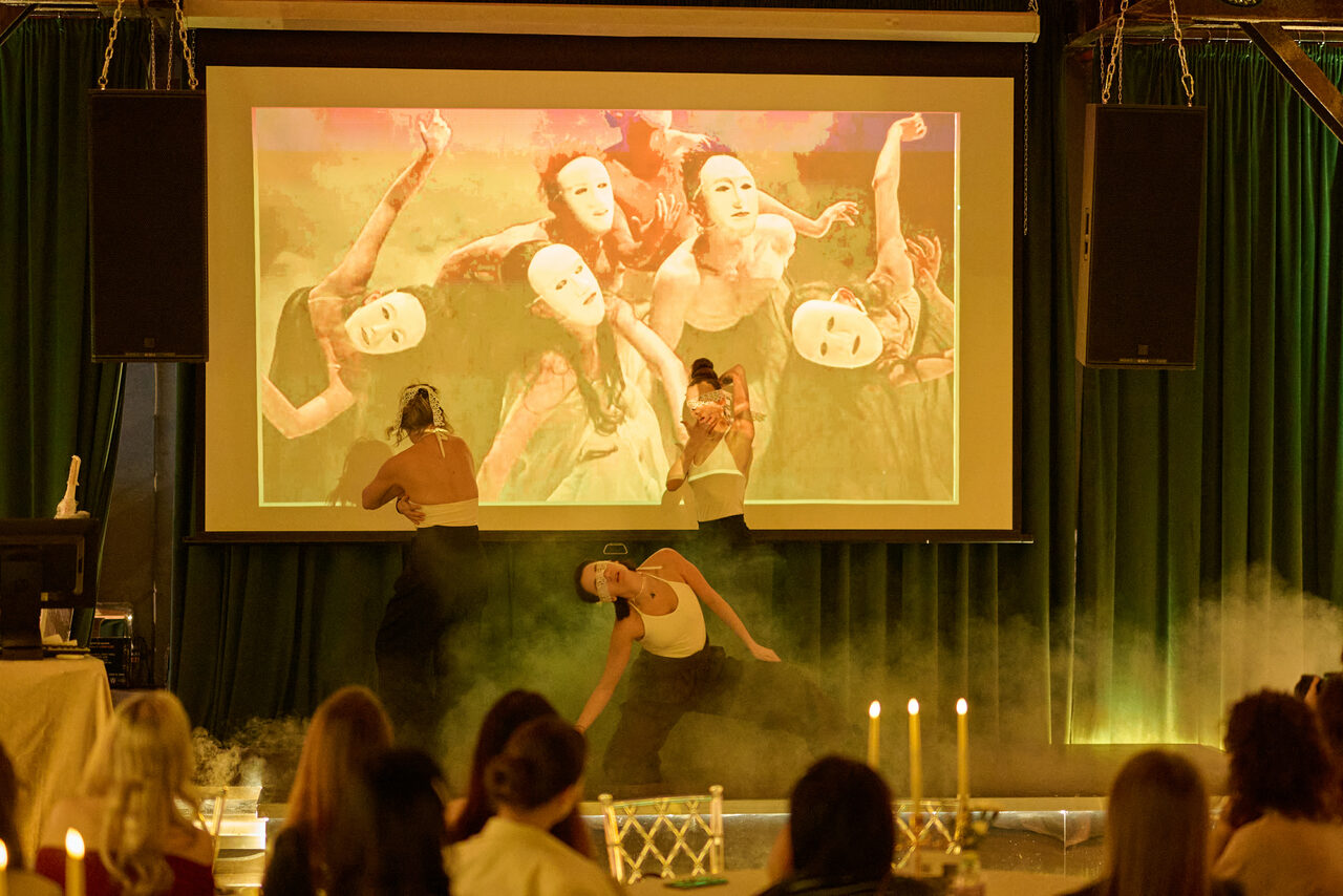 Арендовать Arsenal Loft(Арсенал Лофт). Изображение пространства для мероприятий с проектором, экраном и зеленым фоном. В зале установлены звуковые колонки. Перед сценой сидят люди. На столах стоят свечи в подсвечниках. Освещение тёплое, присутствует дымка.