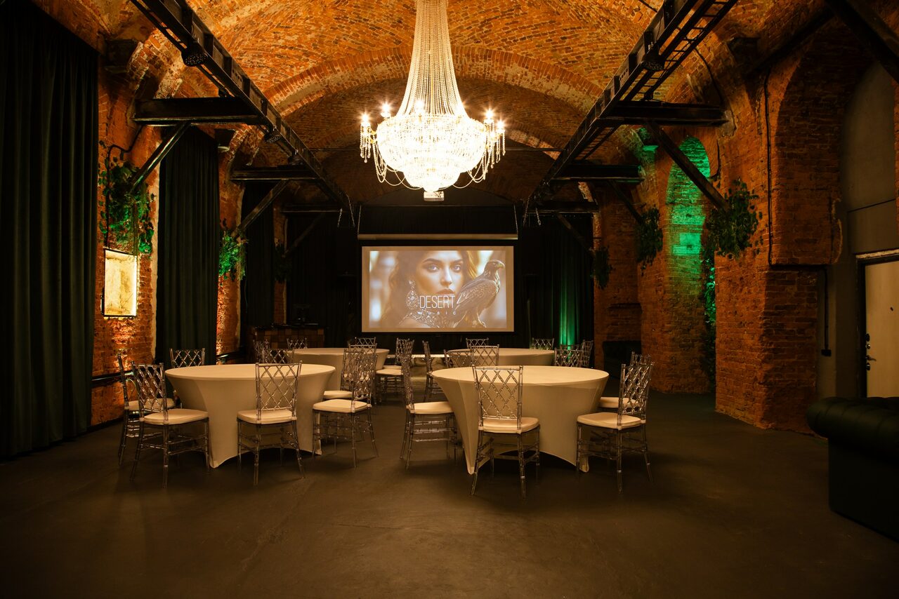 Арендовать Arsenal Loft(Арсенал Лофт). Подвальное пространство с кирпичными арками, круглыми столами и прозрачными стульями. На стене большой экран с изображением. Потолочная люстра освещает помещение. Потолок и стены из красного кирпича, местами зеленые подсветки.