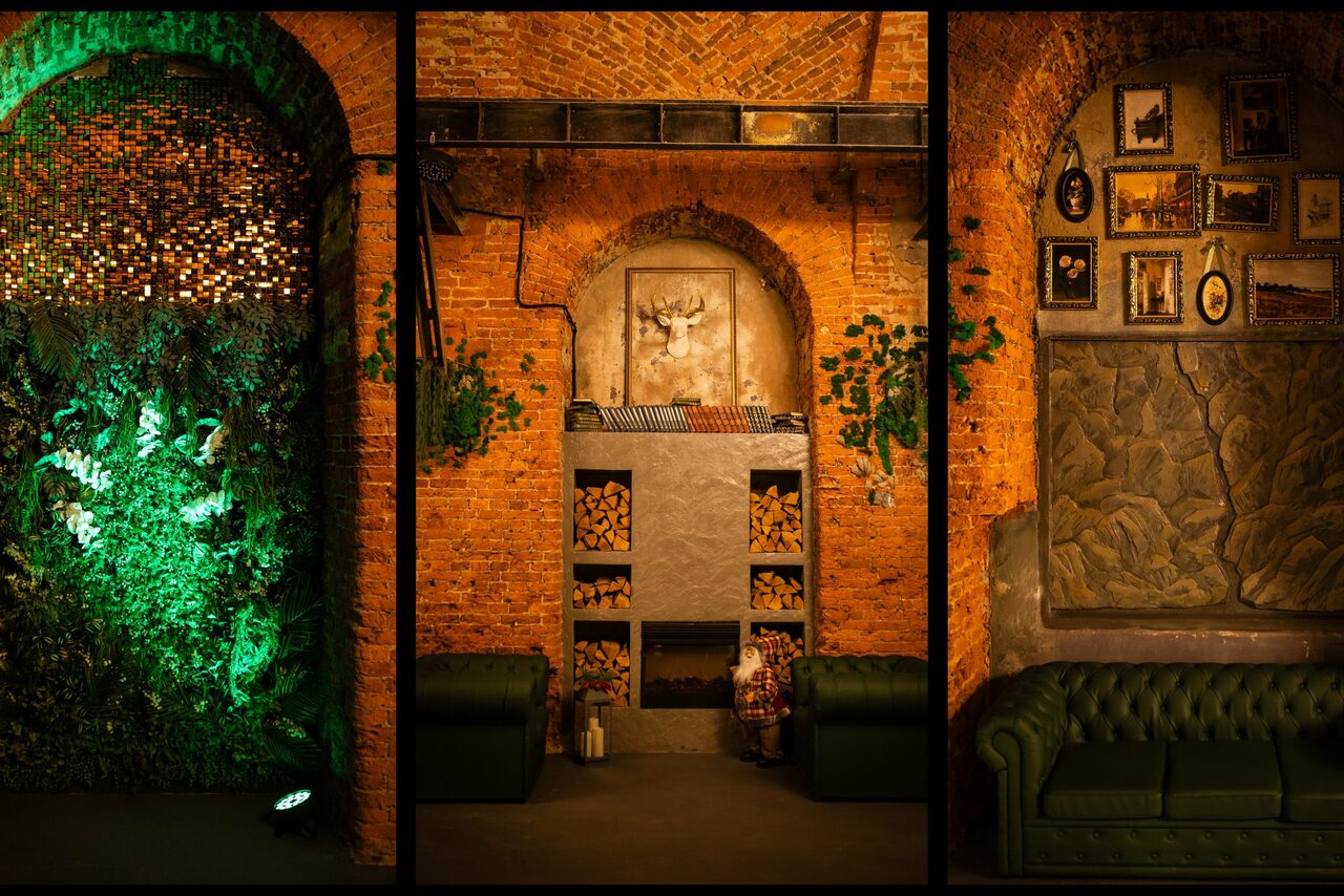 Арендовать Arsenal Loft(Арсенал Лофт). Пространство с кирпичными стенами, разделенное на три зоны. Слева — арка с зелеными растениями и подсветкой. В центре — кладка дров, на стене изображение оленя. Справа — зеленый диван, над ним несколько картин в рамках. Теплое освещение.
