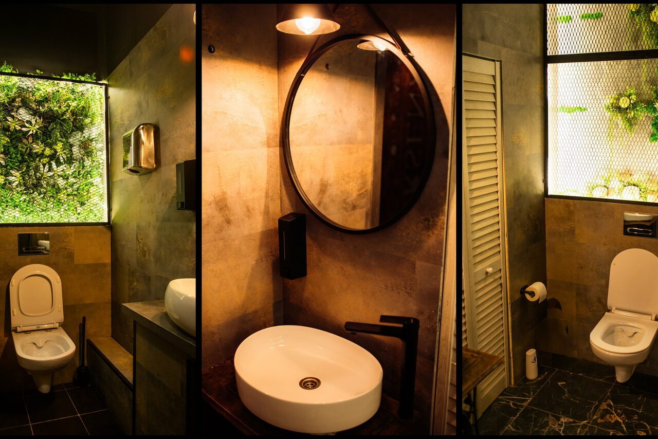 Арендовать Arsenal Loft(Арсенал Лофт). Ванная комната с тремя зонами: унитаз на фоне стены с зелеными растениями, раковина с круглым зеркалом и светильником, второй унитаз и деревянная дверь. Стены и пол отделаны темной плиткой. Освещение теплое, встроенное.