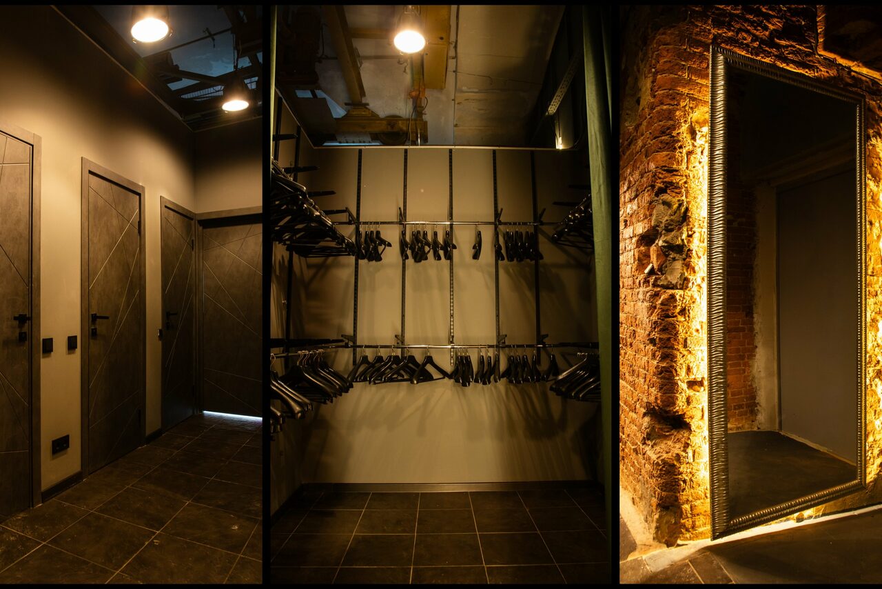 Арендовать Arsenal Loft(Арсенал Лофт). Коридор с тёмной плиткой и дверями, гардеробная зона с вешалками и освещением, кирпичная стена с большим зеркалом в раме. Современное освещение, оттенки серого, коричневого, индустриальный стиль.