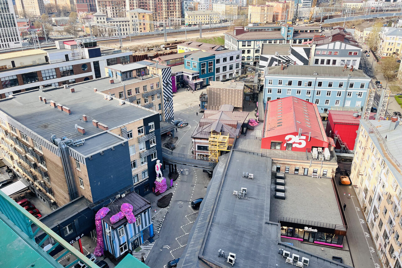 Арендовать Зал под кулинарные мастер классы и мероприятия. Изображение показывает вид на квартал с высоты. Видны несколько зданий разной формы и цвета, в том числе синий, красный и серый фасады. На крыше одного из зданий — большие цифры "25". На улице — розовые скульптуры.