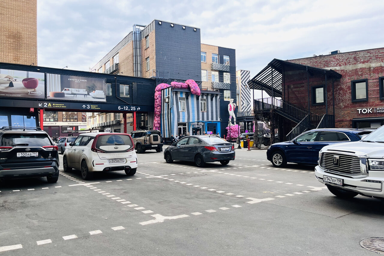 Арендовать Зал под кулинарные мастер классы и мероприятия. Парковка на открытом воздухе с несколькими автомобилями, на заднем плане здания из кирпича и бетона, внешний декор в виде крупного розового цветка, лестница с черным навесом, рекламные билборды, указатели с номерами и логотипами.