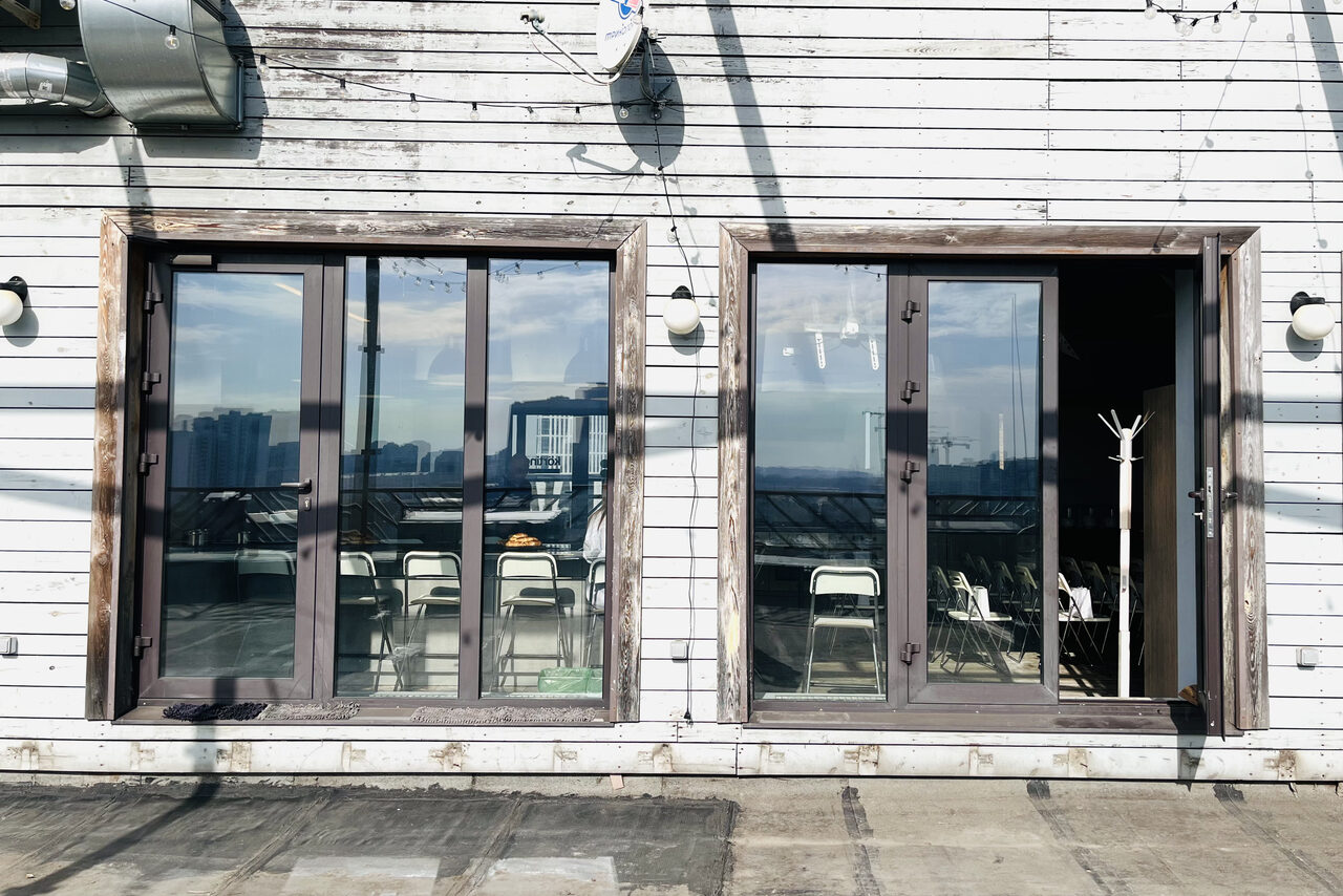 Арендовать Зал под кулинарные мастер классы и мероприятия. Помещение с двумя большими стеклянными дверями на террасе; видно несколько белых стульев и вешалка для одежды внутри. Стены и двери из светлого дерева, снаружи есть светильники. Пол террасы бетонный.