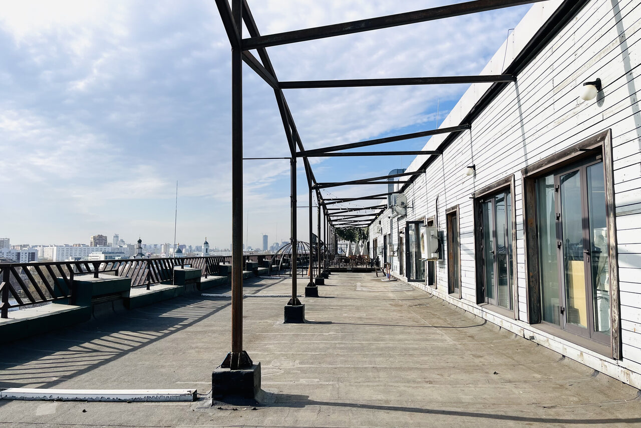 Арендовать Зал под кулинарные мастер классы и мероприятия. Уличная терраса с открытым пространством, металлическими каркасами, деревянными скамейками и перилами, стеклянные двери на заднем плане. Обшивка стен из светлого дерева. Освещение внешнее. Светлый, ясный день.