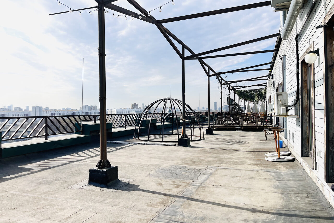 Арендовать Зал под кулинарные мастер классы и мероприятия. Открытая терраса на крыше с бетонным полом, металлическими перголами и ограждением. Справа вдоль стены установлены светильники. По центру пустая металлическая конструкция. На горизонте виден городской пейзаж.