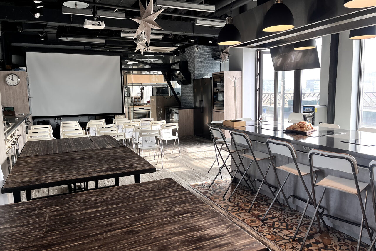Арендовать Зал под кулинарные мастер классы и мероприятия. Пространство с проектором и экраном, множеством белых стульев и деревянными столами. Барная стойка с высокими стульями справа, большие окна, тёмные подвесные светильники, деревянный и плиточный пол. Потолок с открытыми коммуникациями.