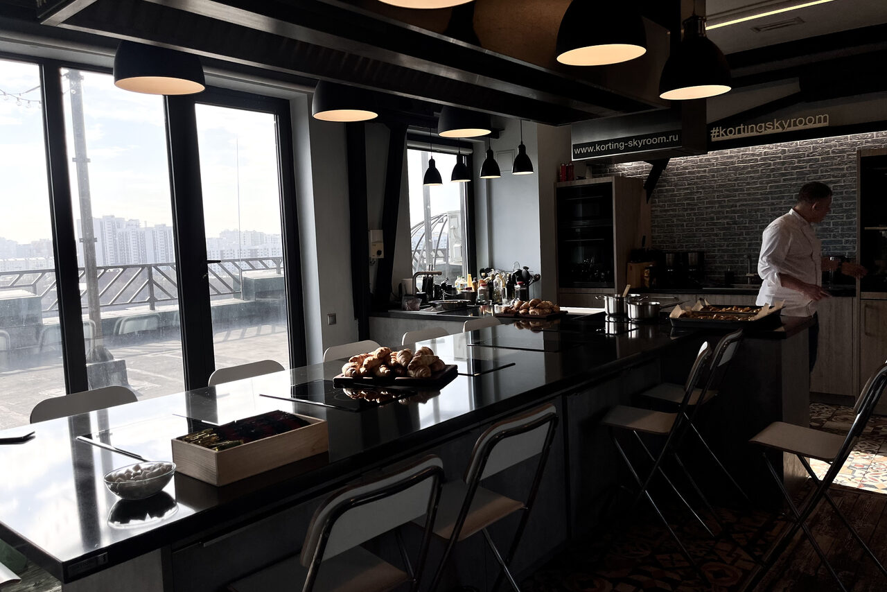 Арендовать Зал под кулинарные мастер классы и мероприятия. Кухонное пространство с длинным столом-островом, черной столешницей, белыми стульями и приборами. Широкие окна с видом на город. Подвесные светильники, кирпичная стена, плитка на полу. На столе булочки и кофеварка.