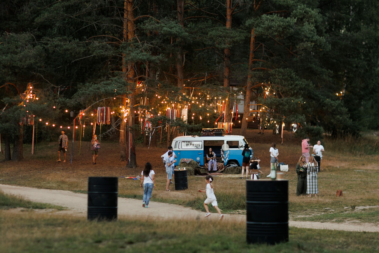 Арендовать Белый Пёс(WHITE DOG event). Лесная поляна с высокими деревьями, украшенная гирляндами. В центре стоит фургон с открытой дверью и людьми вокруг. На земле расставлены большие черные цилиндрические объекты. Видны люди на фоне, бегущие дети.
