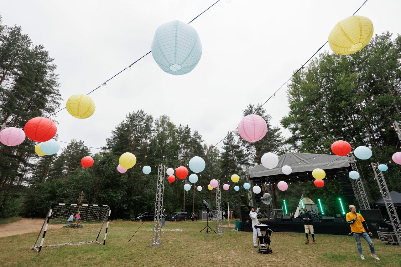 Арендовать Белый Пёс(WHITE DOG event). Открытая площадка на траве, декорированная разноцветными бумажными фонариками, натянутыми на проводах. Сцена со светодиодными экранами и прожекторами. Металлические стойки с колоннами. Футбольные ворота. Лес на заднем плане.