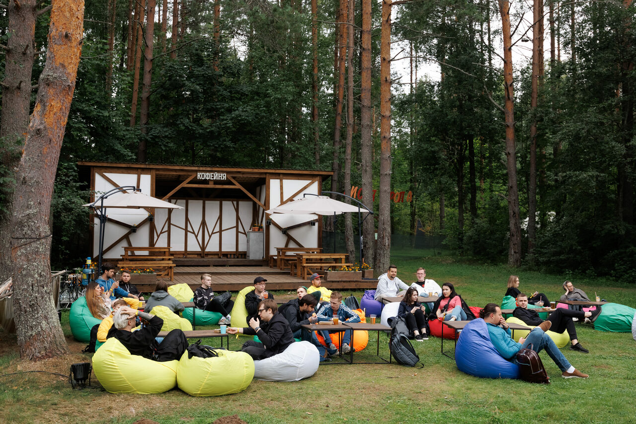Арендовать Белый Пёс(WHITE DOG event). Открытое пространство на траве с пуфами разных цветов и столиками. На фоне небольшая деревянная постройка с надписью. Видны два больших зонта. Вокруг деревья, создающие тень и обрамляющие территорию. Люди сидят, общаются.