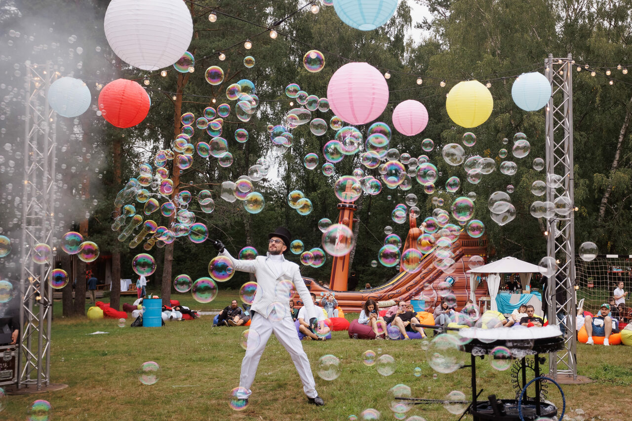 Арендовать Белый Пёс(WHITE DOG event). Открытое пространство с травяным покрытием, подвесные цветные бумажные фонари, металлические конструкции, человек в белом костюме, мыльные пузыри, группа людей сидит на разноцветных креслах-мешках, деревянные детские горки, деревья на заднем плане.