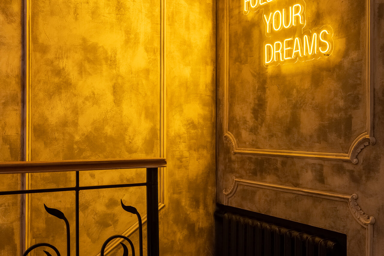 Арендовать Каминный Зал Tangiers lounge Pokrovka(Танжерс Лаундж Покровка). Золотистая стена с декоративными элементами, черный радиатор. Металлические перила слева внизу. Яркая неоновая надпись на английском языке. Теплое освещение.