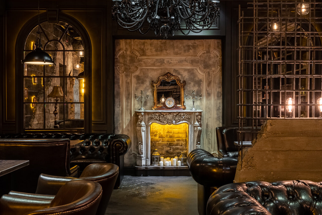 Арендовать Каминный Зал Tangiers lounge Pokrovka(Танжерс Лаундж Покровка). Интерьер в стиле лофт с кожаными креслами и диванами, декоративным камином и зеркалом, свечи в камине, черная люстра, арочные окна, металлическая решётка, кирпичная стена, тёплое освещение.