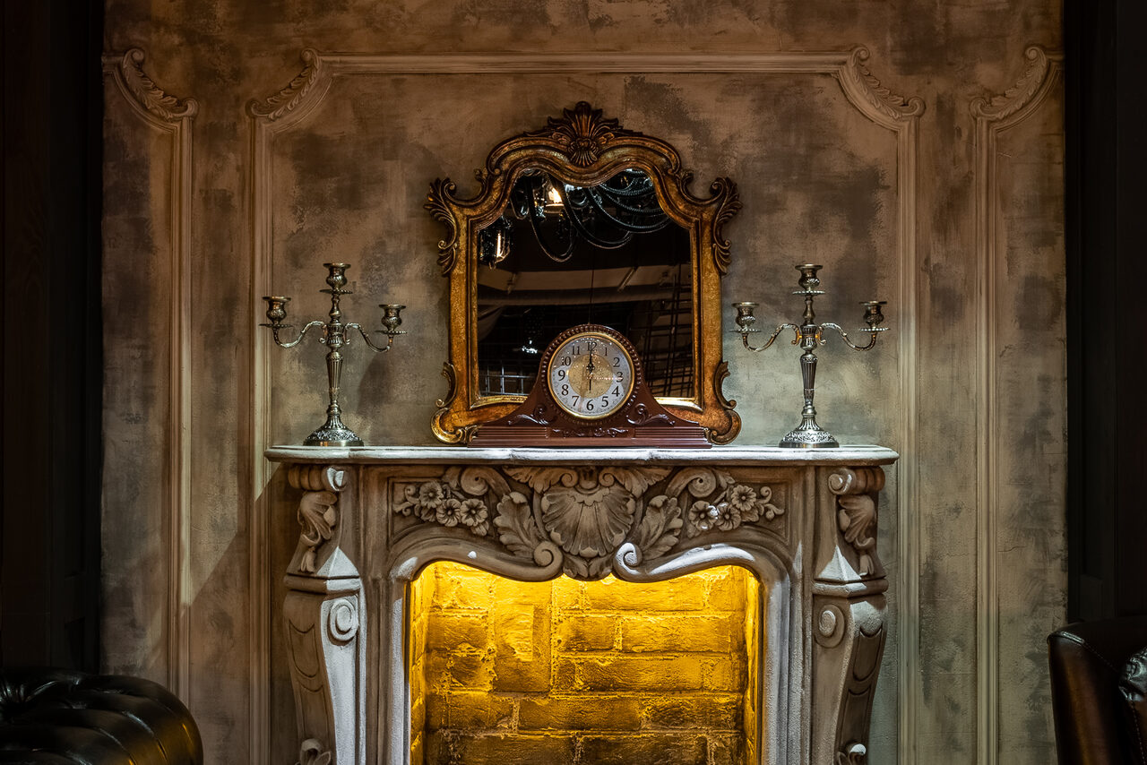 Арендовать Каминный Зал Tangiers lounge Pokrovka(Танжерс Лаундж Покровка). Камин с резным декором и подсветкой, на каминной полке зеркало в резной раме, два металлических подсвечника и настольные часы. Стена с декоративной отделкой, коричневые и золотистые оттенки.