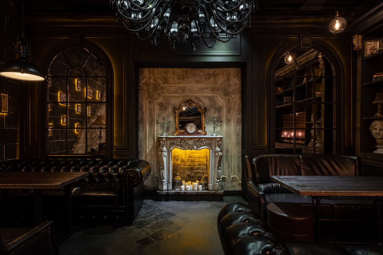 Арендовать Каминный Зал Tangiers lounge Pokrovka(Танжерс Лаундж Покровка). Тёмное лаунж-пространство с кожаными диванами и деревянными столами. Центрально располагается декоративный камин с зеркалом и свечами. Арочные окна, полки с книгами, бюст. Освещение жёлтое, мягкое, с крупной чёрной люстрой.