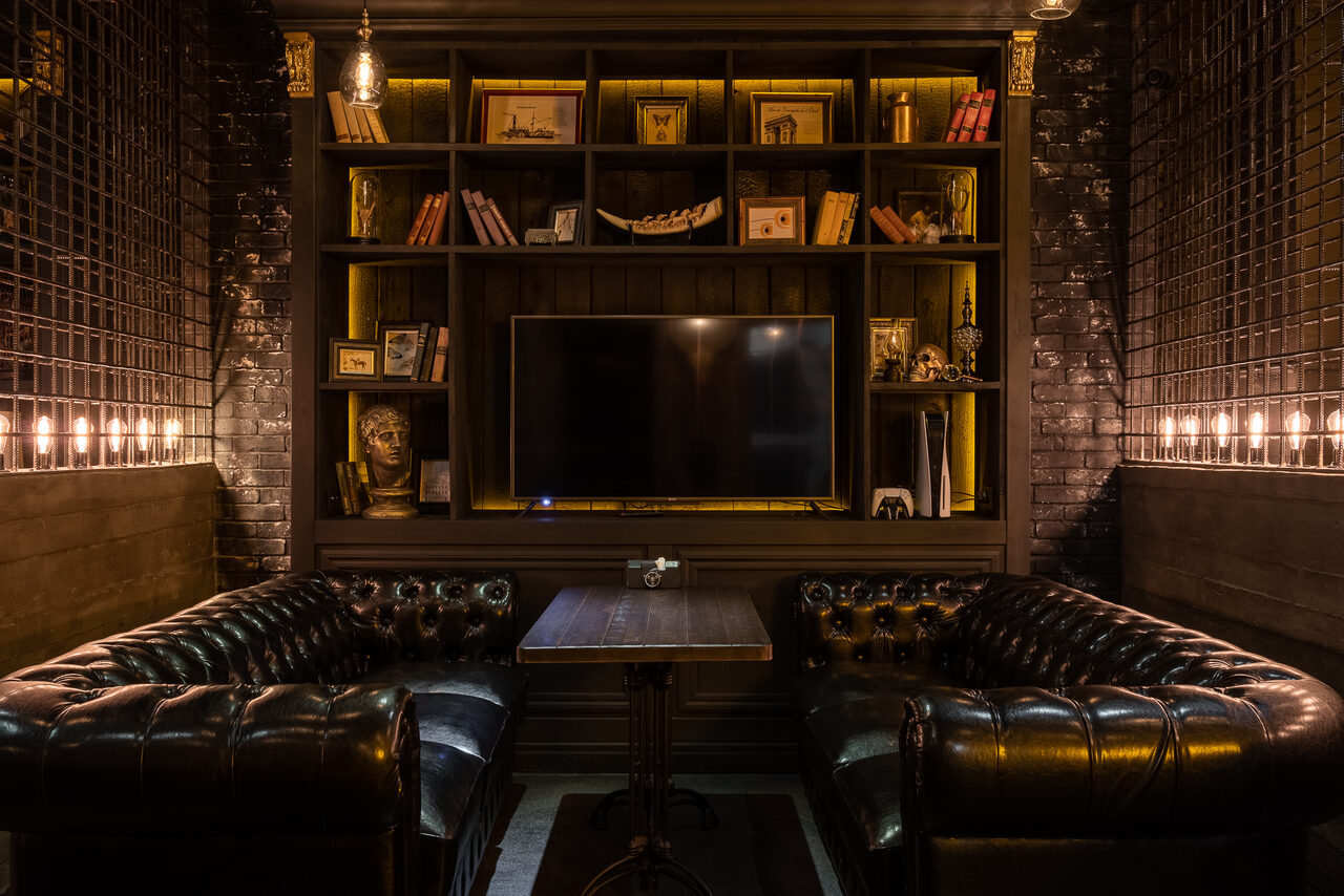 Арендовать Каминный Зал Tangiers lounge Pokrovka(Танжерс Лаундж Покровка). Пространство с двумя кожаными диванами и деревянным столом. Стена с кирпичной кладкой, встроенные полки с книгами и декором. Большой телевизор по центру. Теплое освещение с лампами. Темные тона интерьера.