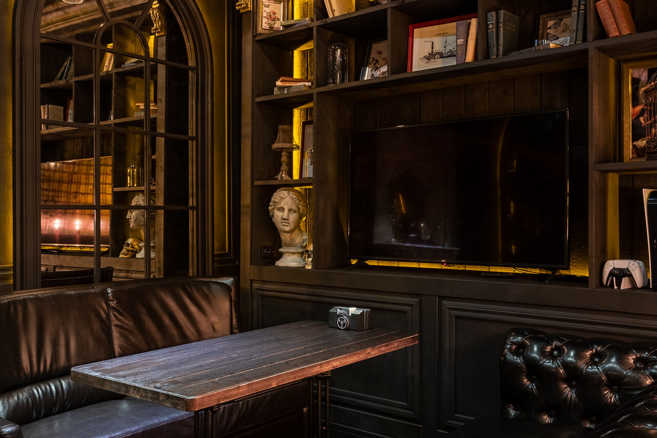 Арендовать Каминный Зал Tangiers lounge Pokrovka(Танжерс Лаундж Покровка). Частная комната с кожаным диваном, деревянным столом, книжными полками и телевизором на стене. Стены темные, внутреннее освещение теплое. На полках стоят книги, декоративные элементы и статуэтка. На фоне зеркало в раме.