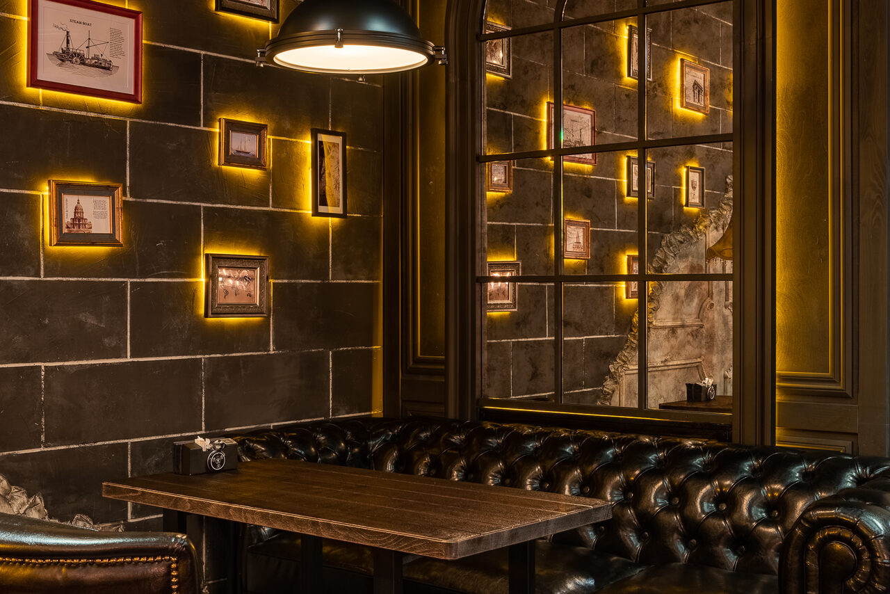 Арендовать Каминный Зал Tangiers lounge Pokrovka(Танжерс Лаундж Покровка). Интерьер кафе или бистро с кожаными диванами, деревянным столом, кирпичными стенами с подсветкой и фотографиями в рамах. Освещение теплое, подвесной светильник над столом. Стены темного цвета, декоративные элементы создают уютную атмосферу.