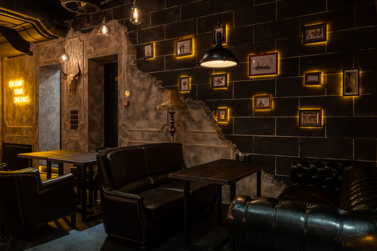 Арендовать Каминный Зал Tangiers lounge Pokrovka(Танжерс Лаундж Покровка). Лофт-пространство с кожаными диванами и деревянными столами. Стены оформлены в индустриальном стиле с чёрными панелями и светящимися рамками с небольшими изображениями. Потолочные и настенные светильники, тёплое освещение. Текст: "FOLLOW YOUR DREAMS".