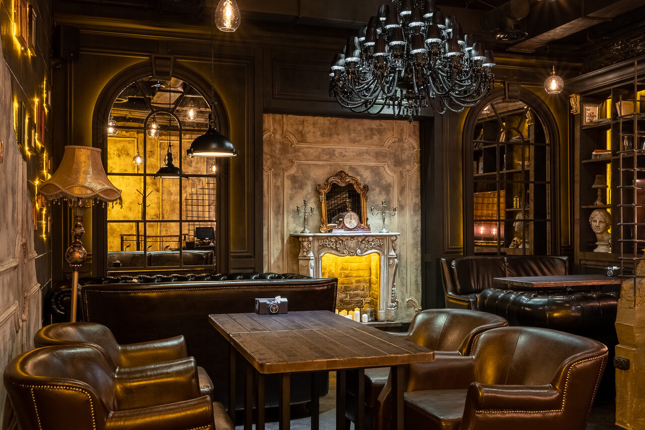 Арендовать Каминный Зал Tangiers lounge Pokrovka(Танжерс Лаундж Покровка). Просторное помещение с темной деревянной мебелью, кожаными креслами и диванами. Центральное место занимает камин. Над ним большое зеркало в раме. Потолочный черный канделябр и настенные светильники создают теплый свет. На стенах висят зеркала.