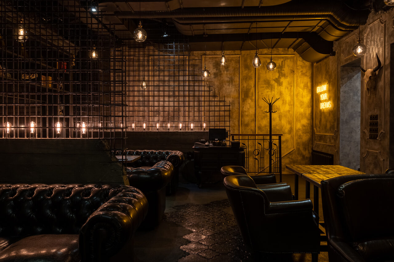 Арендовать Каминный Зал Tangiers lounge Pokrovka(Танжерс Лаундж Покровка). Тёмное пространство с элементами индустриального стиля. В помещении кожаные диваны, деревянный стол, металлическая сетка, подвесные лампы. На стене неоновая надпись. Пол светло-серый, стены покрыты штукатуркой. Освещение теплое.