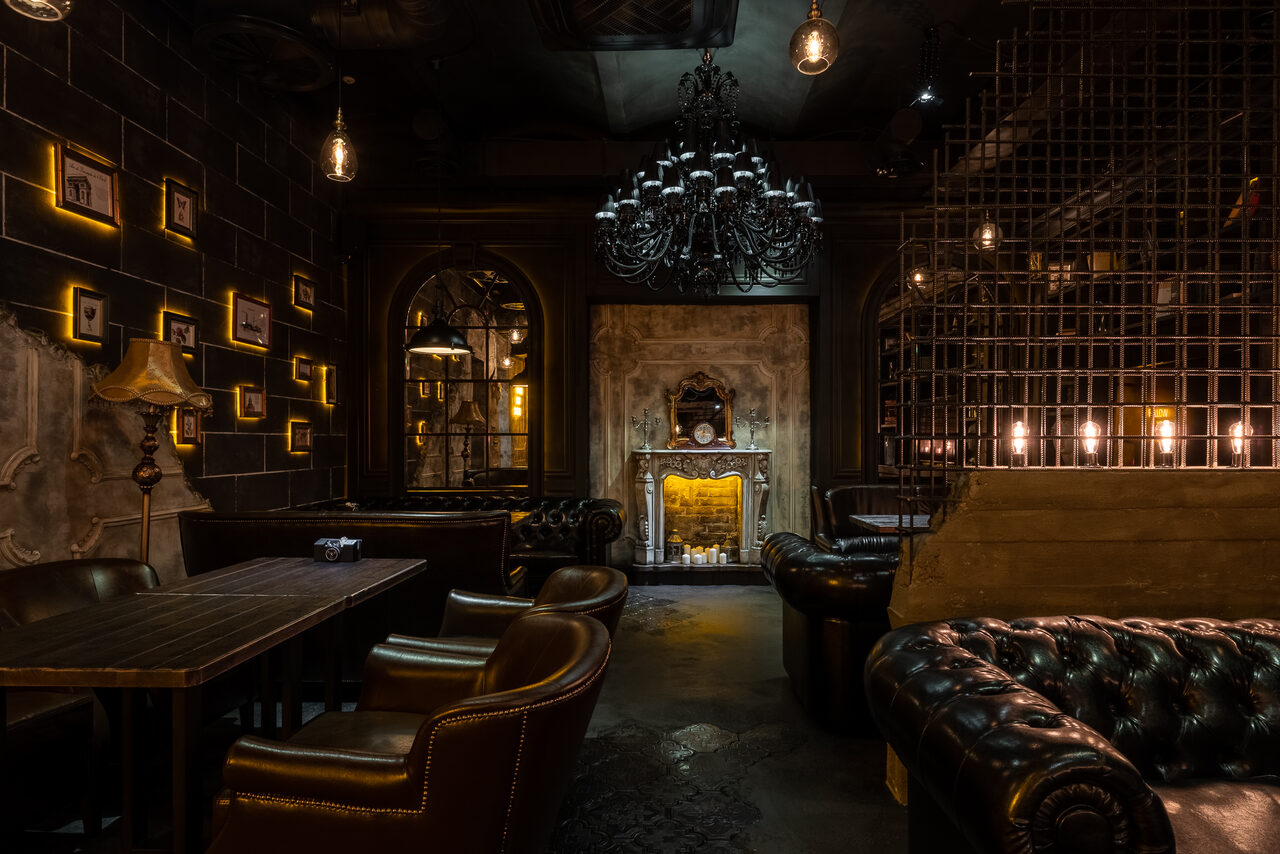 Арендовать Каминный Зал Tangiers lounge Pokrovka(Танжерс Лаундж Покровка). Тёмное и атмосферное пространство с кожаными диванами и креслами, деревянными столами, декоративным камином. Освещение обеспечивают подвесные лампы и свечи. Стены оформлены фоторамками и светильниками. Металлические решётки в интерьере.