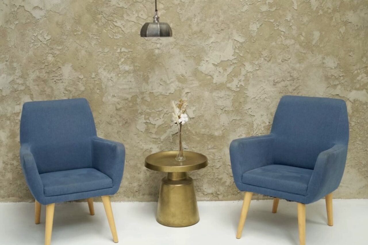 Арендовать WhiteFox Studio(Студия Белая Лиса). Две синие кресла с деревянными ножками стоят по бокам круглого золотистого столика с вазой на фоне декоративной стены с фактурной отделкой. Над столиком подвешен металлический светильник. Пол светлый.