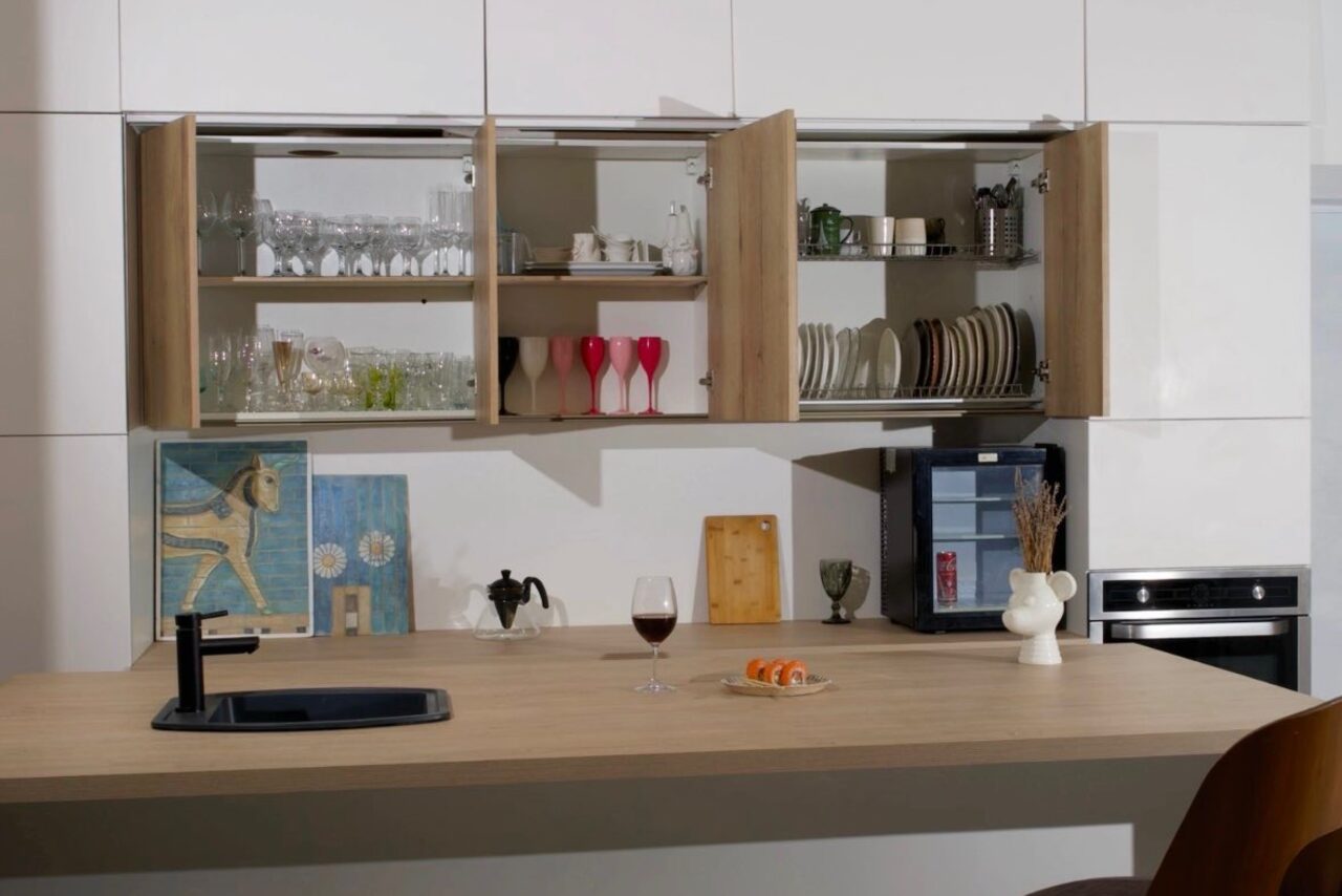 Арендовать WhiteFox Studio(Студия Белая Лиса). Кухонное пространство с деревянной столешницей, открытые шкафы со стеклянной и керамической посудой, картина, ваза на столе, черный кран, бокал с напитком, тарелка с едой, встроенная духовка, светодиодное освещение, нейтральные оттенки.