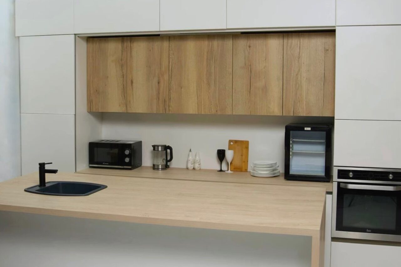 Арендовать WhiteFox Studio(Студия Белая Лиса). Современная кухня с деревянной столешницей и шкафами. Есть встроенная духовка, микроволновка, чайник, раковина с краном, холодильник, стеклянные бокалы, стопка тарелок, разделочная доска. Декор светлый, с деревянными и черными акцентами.