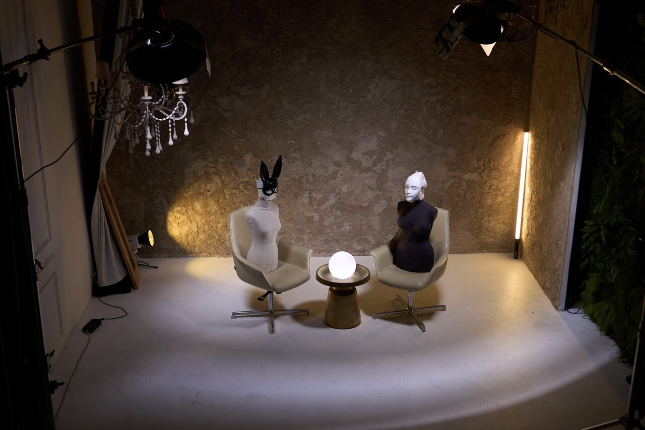 Арендовать WhiteFox Studio(Студия Белая Лиса). Стильное пространство с двумя креслами и маленьким столиком посередине. На креслах размещены манекены. На фоне фактурная стена и вертикальный светильник. Освещение мягкое, дополнительный свет от потолочных ламп. Полы светлые.