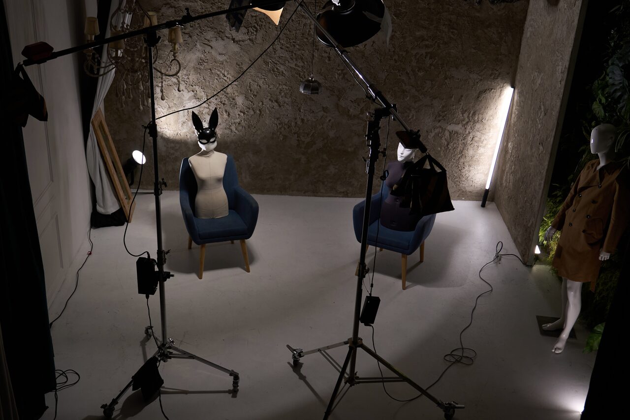Арендовать WhiteFox Studio(Студия Белая Лиса). Студийное пространство с двумя синими креслами, манекенами в масках, прожекторами на штативах, светодиодными светильниками вдоль стен. Стены из грубого серого материала, пол светлый. На заднем плане манекен в коричневом пальто.