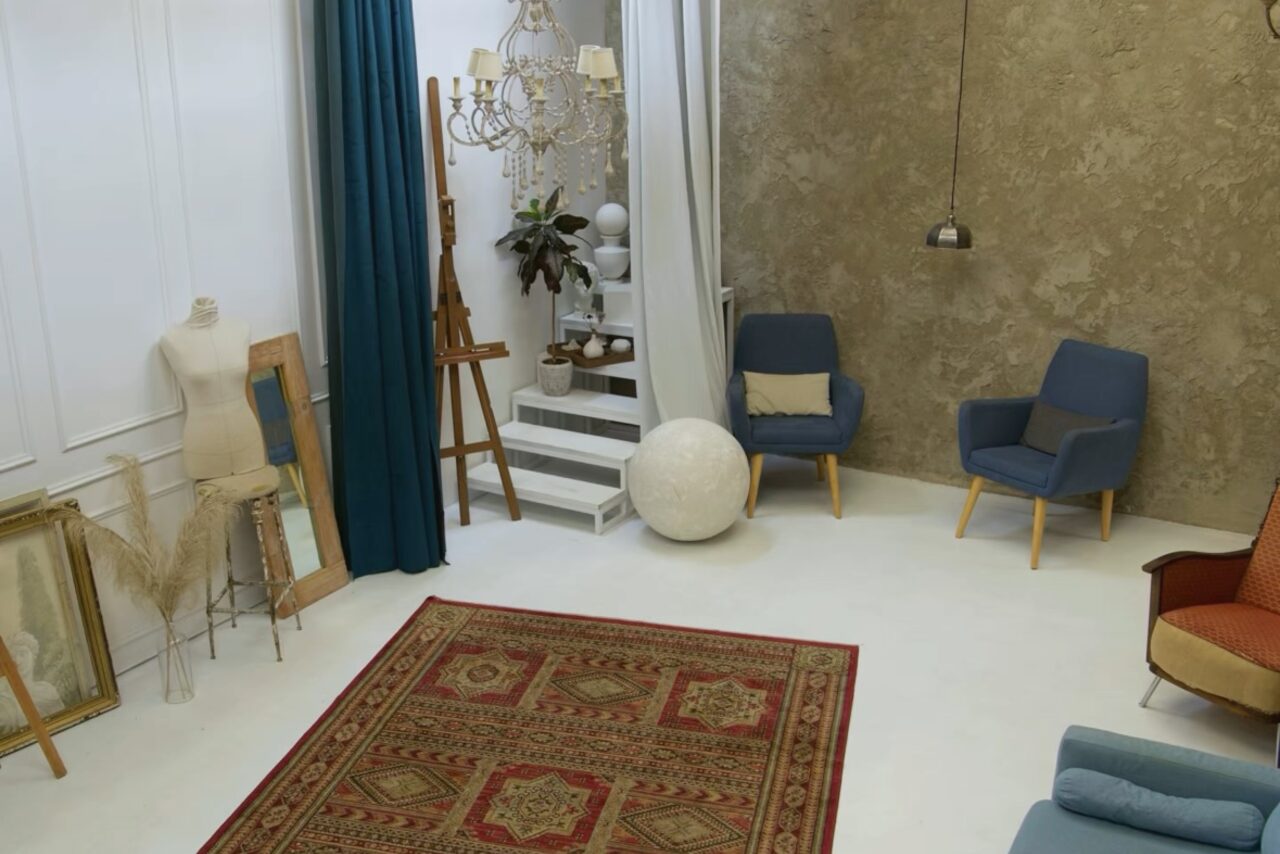 Арендовать WhiteFox Studio(Студия Белая Лиса). Просторная комната с белыми стенами и полом. Два синих кресла, коричневое кресло и манекен. Настенные украшения, большая лампа, зеленая штора и декоративная лестница. Пёстрый ковер на полу, подвесной светильник.