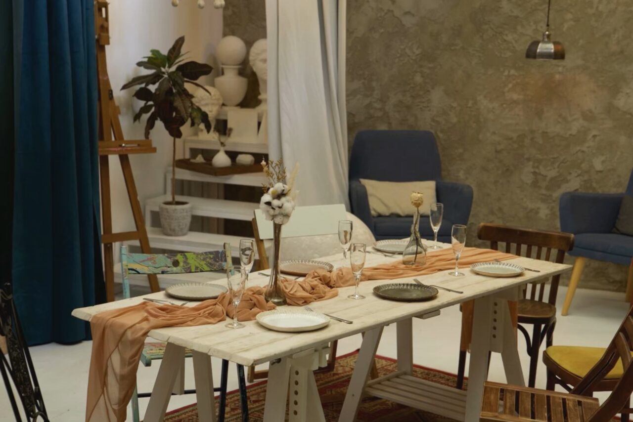 Арендовать WhiteFox Studio(Студия Белая Лиса). Столовая зона с белым деревянным столом, сервированным тарелками и бокалами. Окружена разными стульями. На фоне серая фактурная стена, синие шторы, полки с растениями и скульптурами. Освещение висит сверху. Теплые и нейтральные цвета.