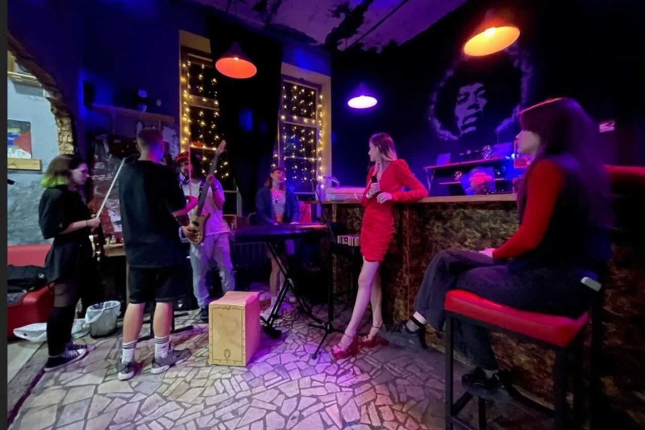 Арендовать Лофт-Бар. Музыкальное пространство с барной стойкой, на стене портрет музыканта. Группа людей с инструментами, включая клавиши и саксофон. Красные и фиолетовые подвесные светильники, фонари за окном. Плиточный пол, разноцветная мебель.