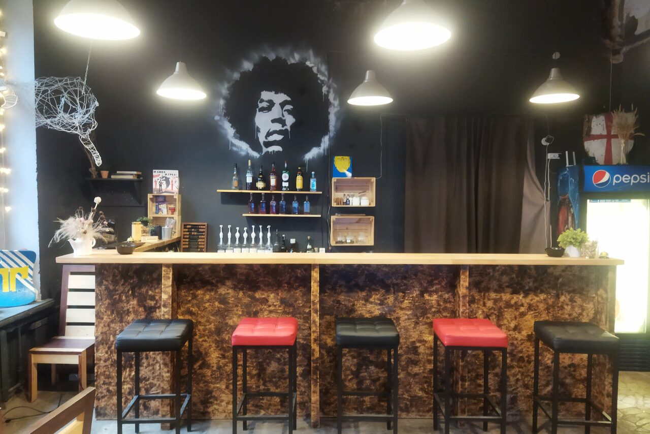 Арендовать Лофт-Бар. Барная стойка с пятью стульями, чередующимися чёрными и красными сиденьями. На темной стене висит изображение человека. Справа холодильник для напитков. Освещение представлено подвесными светильниками. На стойке и полках алкогольные бутылки.