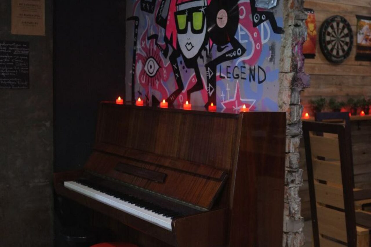 Арендовать Лофт-Бар. Кафе или бар с угловым расположением настенного пианино, рядом рисунок граффити на стене. На пианино стоят небольшие свечи. На стене справа дартс и деревянная обшивка. Оттенки коричневого, красного, фиолетового и синего.