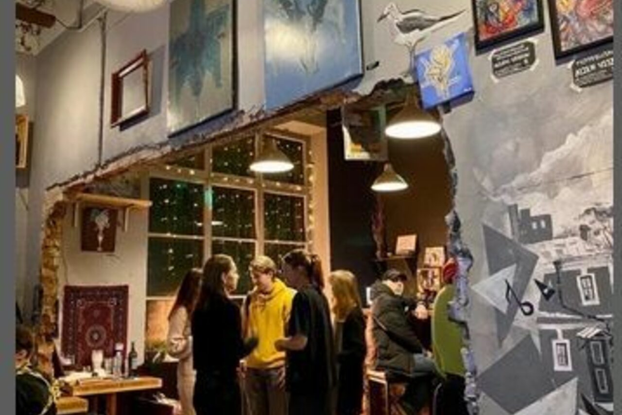 Арендовать Лофт-Бар. Творческое пространство с арт-декором, картинами на стенах, открытая кирпичная кладка, подвесные светильники, винные бутылки на столах, люди общаются, деревянная мебель, большая декорированная стена с рисунками, большое окно с гирляндами за стеклом.