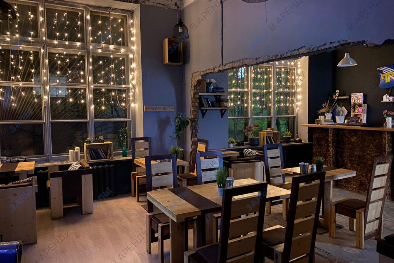 Арендовать Лофт-Бар. Пространство в стиле кафе с деревянными столами и стульями, декорированное гирляндами на окнах. Барная стойка слева, небольшие растения на полках и столах, мягкое рассеянное освещение. стены тёмно-серого цвета; пол светлый.