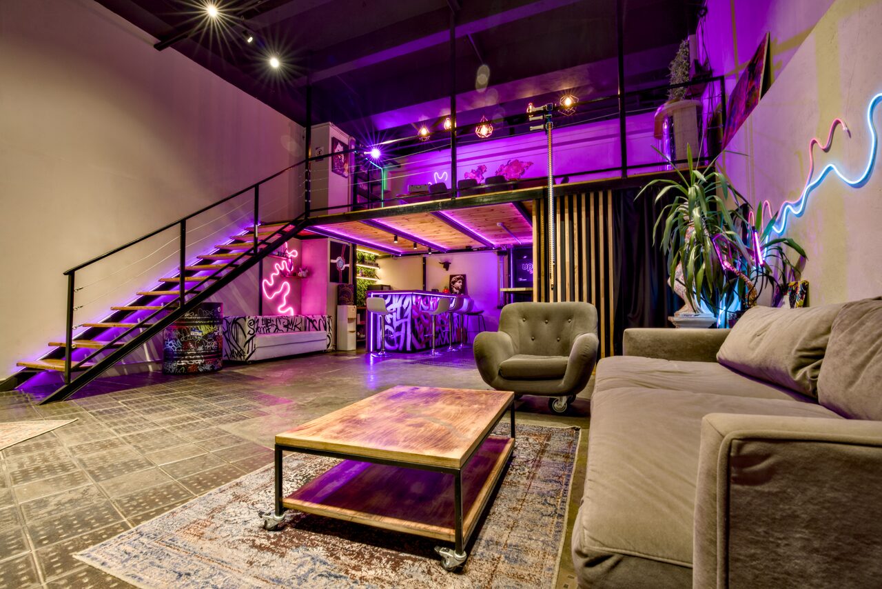Арендовать НЕОН Лофт. Лофт-пространство с мезонином, металлической лестницей, фиолетовой подсветкой. На первом уровне серый диван, кресло, деревянный стол. На втором уровне барная зона. Зелёные растения и неоновая надпись на стене. Тёмный потолок и пол.