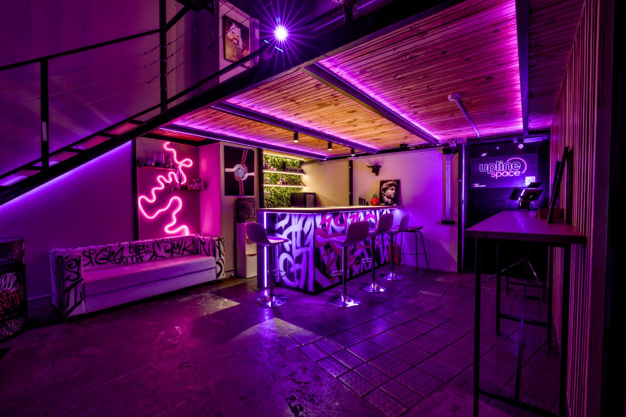 Арендовать НЕОН Лофт. Барное пространство с неоновым освещением, лиловыми акцентами. Барная стойка украшена граффити, несколько высоких стульев. Слева белый диван, на стене неоновый знак, пол бетонный, потолок деревянный. Видна лестница на второй уровень.