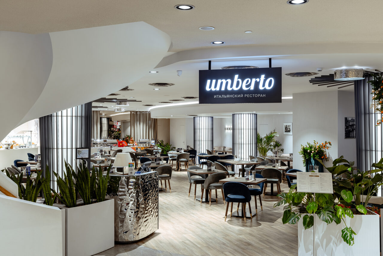 Арендовать Итальянский ресторан Umberto(Умберто). Современный ресторан с открытой планировкой, круглые столы и мягкие стулья, растения в белых горшках, светлые деревянные полы, круглые потолочные светильники, металлическая барная стойка, черный подвесной знак с надписью "умберто итальянский ресторан".