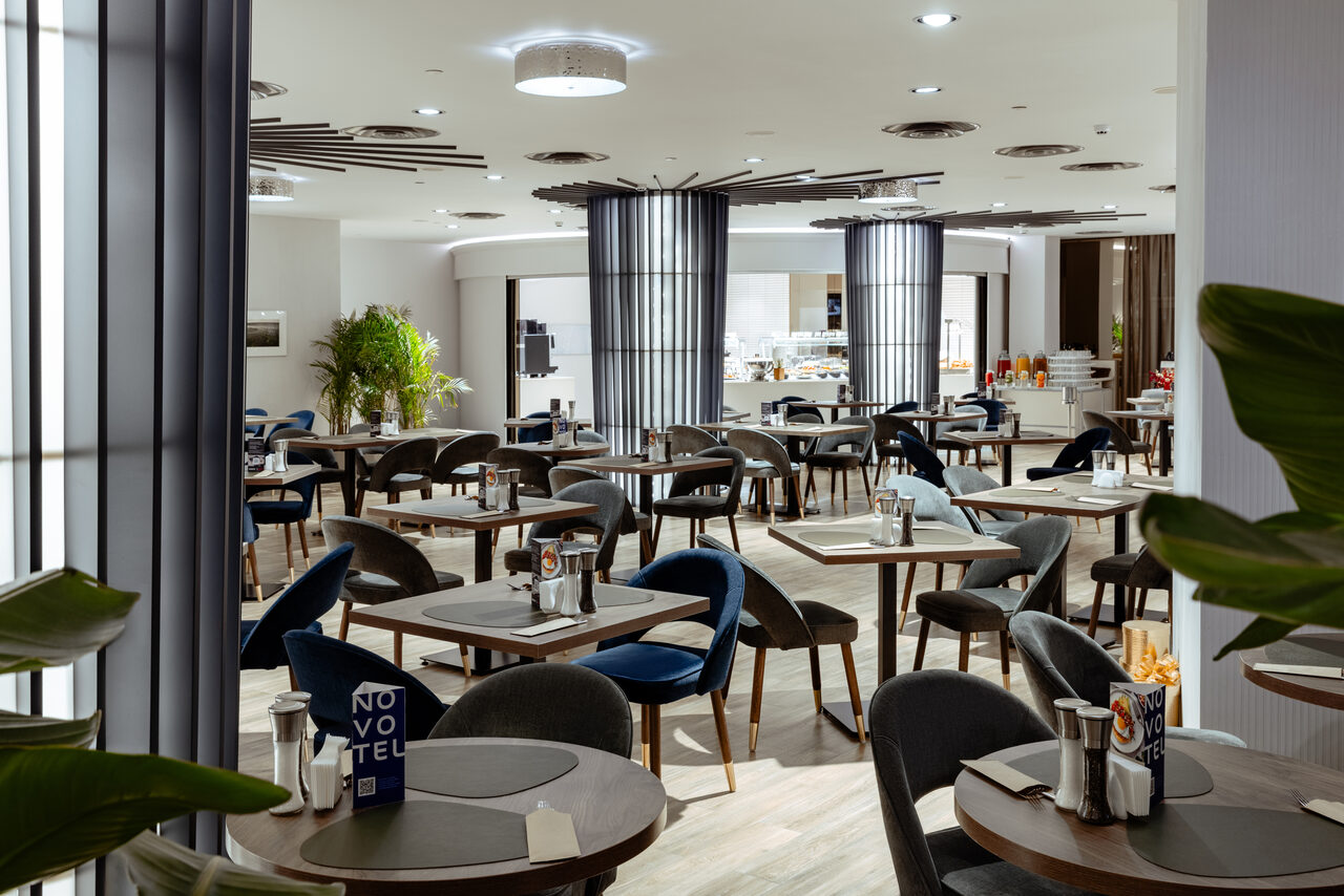 Арендовать Итальянский ресторан Umberto(Умберто). Современный ресторан с круглыми и квадратными столами, темно-синими стульями, мягким освещением. Светлая отделка, деревянные полы, декоративные растения. Пространство для фуршета с напитками и закусками на фоне, надпись "NOVOTEL" на столах.