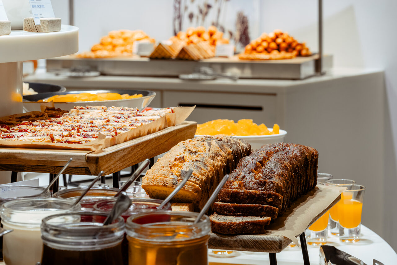 Арендовать Итальянский ресторан Umberto(Умберто). Стол с разнообразными десертами: пироги, кексы и хлеб на подносах, банки с джемом; задний план — апельсиновые шарики и сочные кусочки фруктов. Светлое помещение с нейтральной цветовой палитрой.