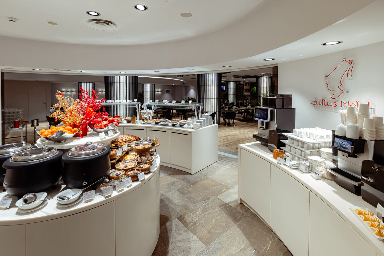 Арендовать Итальянский ресторан Umberto(Умберто). Современное кафе с круглой барной стойкой, сервированной выпечкой и фруктами. Светлые стены, стеклянные дисплеи, кофемашина, посуда. Освещение встроенными светильниками. На стене световой декор. Пол из светлой плитки.