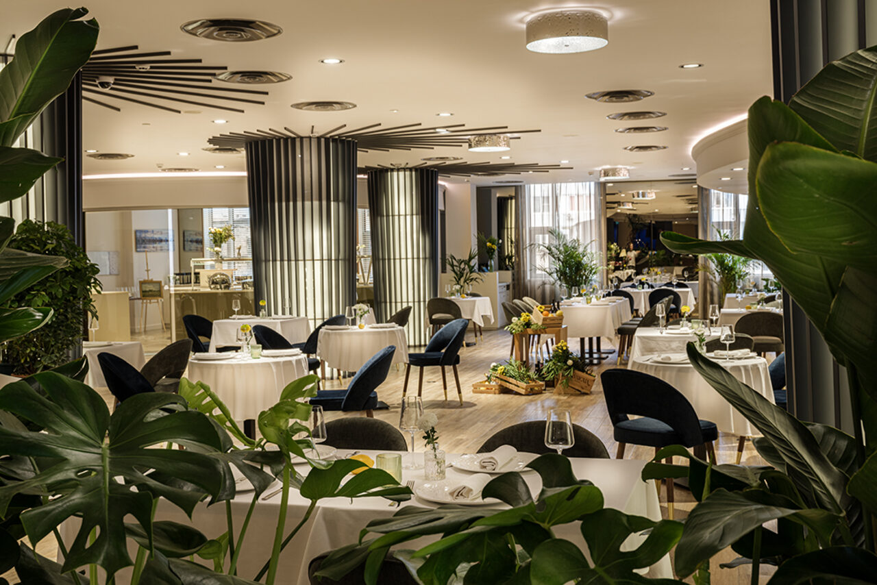 Арендовать Итальянский ресторан Umberto(Умберто). Пространство ресторана с круглым столами, накрытыми белыми скатертями, темными стульями, зелеными растениями. Светлые стены, подвесные потолочные светильники, деревянный пол. Декор из листьев на первом плане.